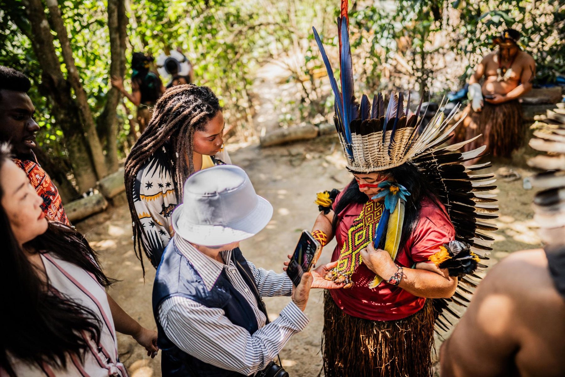 A woman in a tall feathered headdress and red shirt shows beaded jewelry to visitors in a sunlit forest clearing