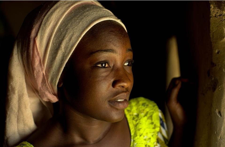 A young woman in a loosely wrapped headscarf looks out of a dark doorway, her face lit by a single stream of light
