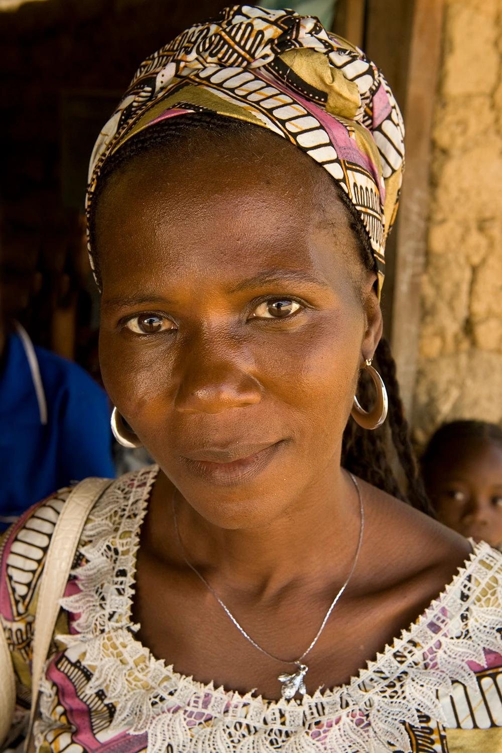 Close portrait of a smiling woman in a patterned headwrap and lace-trimmed top, gold hoop earrings, standing in a doorway