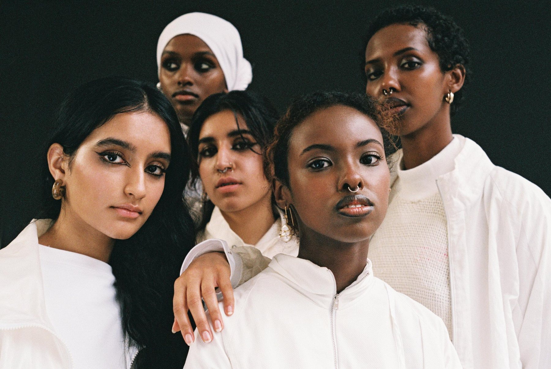 Five women in white clothing against a black backdrop, gathered close and looking straight at the camera